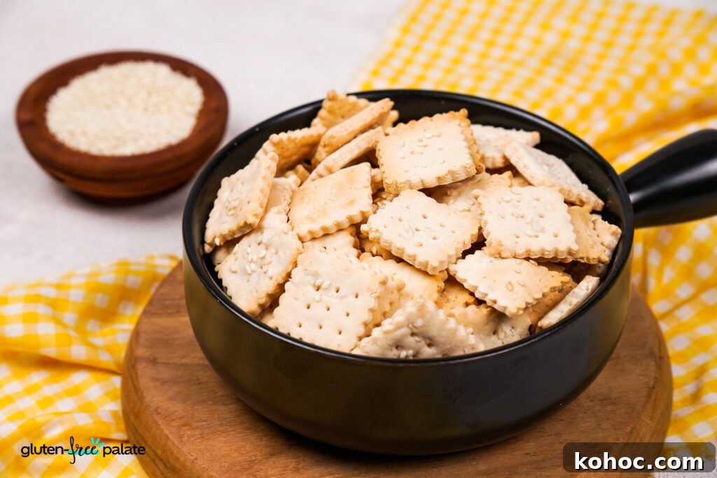 A black bowl filled with square, golden-brown gluten-free crackers topped with sesame seeds, placed on a wooden board with a yellow checkered cloth and a small wooden bowl of sesame seeds in the background.