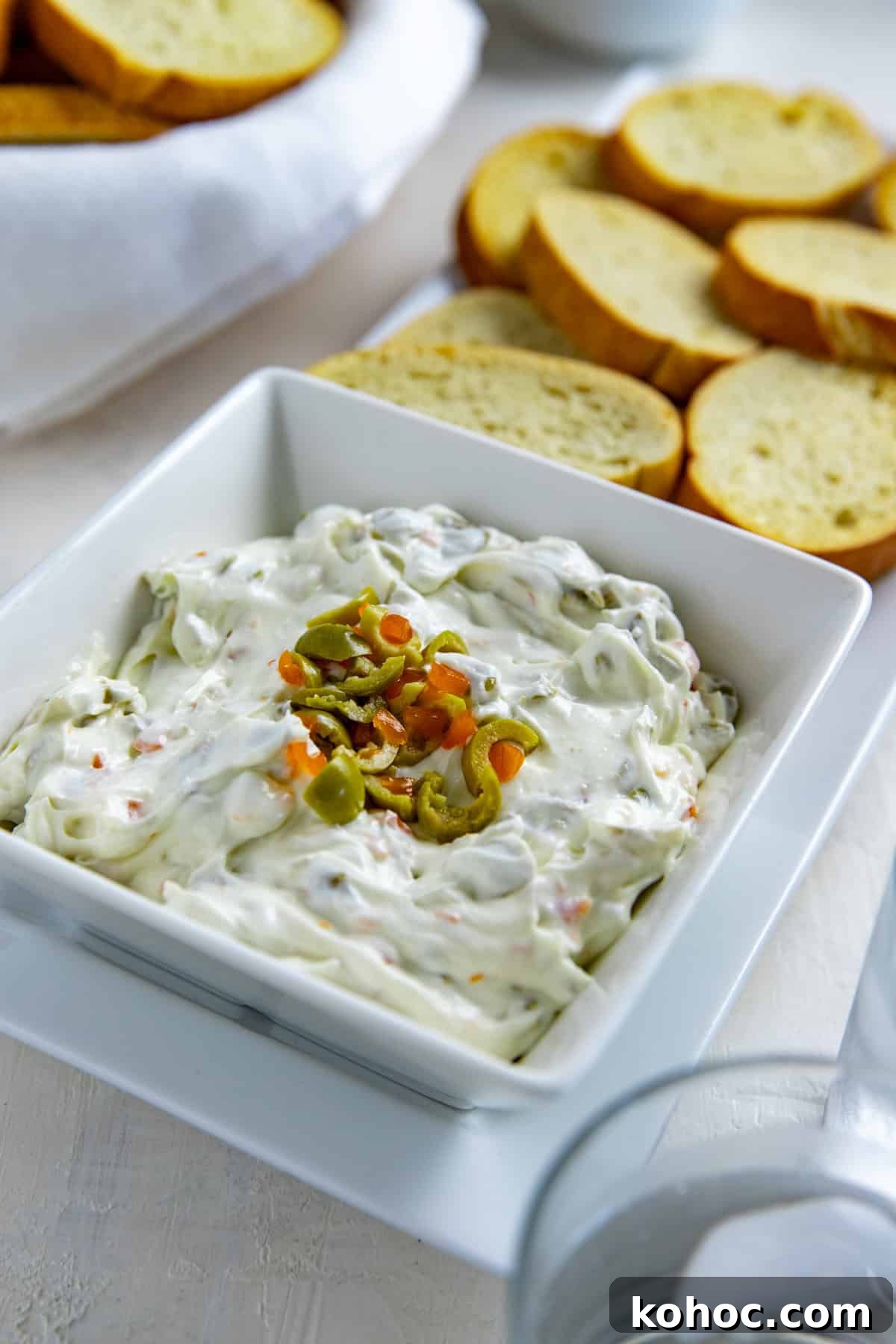 Olive dip in a square, white serving bowl topped with chopped green olives. Crostini in the background.