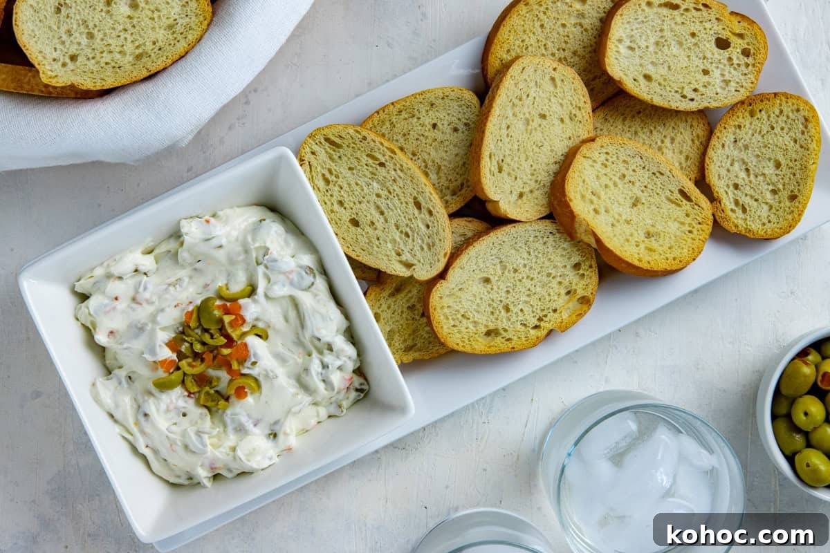 Olive dip in a square, white serving bowl topped with chopped green olives. Crostini in the background.