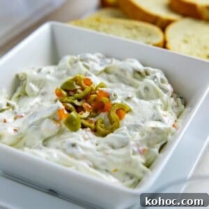 Olive dip in a square, white serving bowl topped with chopped green olives. Crostini in the background.