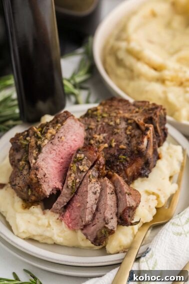 Sliced medium-rare lamb loin chops served on a bed of mashed potatoes, garnished with herbs, with a fork on the plate and a bowl of mashed potatoes in the background.